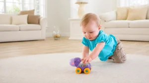 Imagem de bebê brincando com um brinquedo de empurrar roxo e amarelo no tapete da sala, com decoração clara e sofá ao fundo.