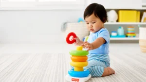 Bebê sentado no tapete branco brincando com um brinquedo empilhável de anéis coloridos e peças em forma de torre, em ambiente claro com prateleira ao fundo.