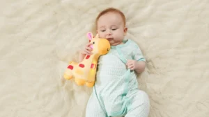 Imagem de bebê de um ano sorrindo enquanto segura um brinquedo de pelúcia em forma de girafa, com roupa azul clara e fundo bege de tecido macio.