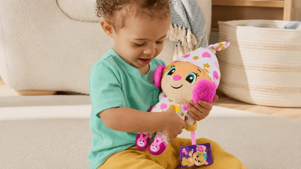 Imagem de criança sentada no chão brincando com um brinquedo de pelúcia colorido em formato de unicórnio, com alça e detalhes de estrelas e coração.