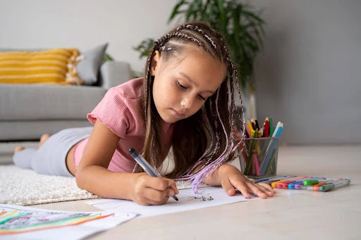 Imagem de criança desenhando em uma mesa com lápis de cor e canetinhas, concentrada e sorrindo, em um ambiente de sala de estar com almofadas e materiais escolares ao fundo