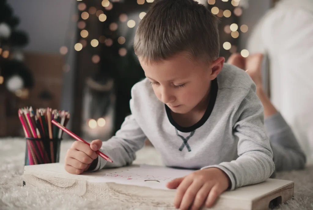 Imagem de criança desenhando com lápis sobre uma mesa, em ambiente aconchegante com luzes ao fundo, usando pijama claro e concentrada na atividade artística.